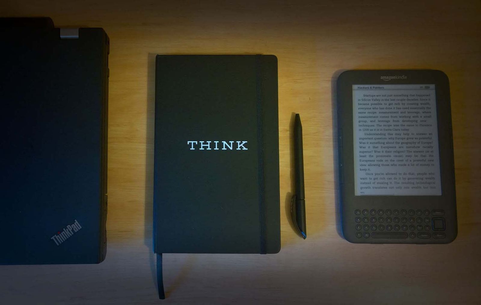 A ThinkPad, a black notebook embossed THINK, a pen and a Kindle on a wooden desk.